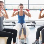 Three women are sitting on a ball and holding blue dumbbells.